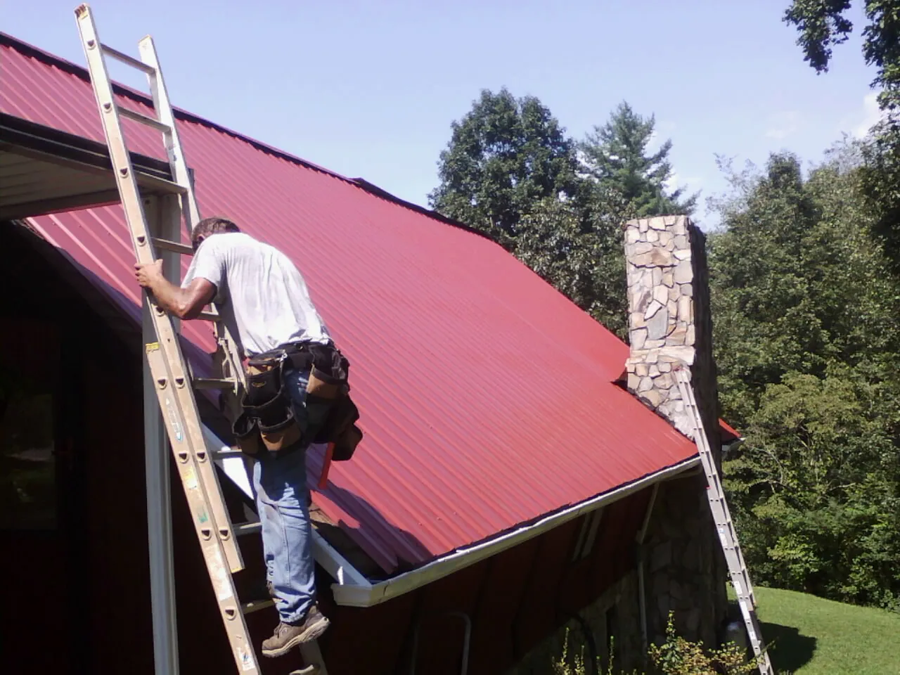 Roofer on a ladder beside a red metal roof and stone chimney — Soffit & Fascia Repair by Roofing Brothers in Mutual